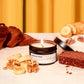 Candle labeled 'Banana Nut Bread' with banana, bread, and nuts on a table against a yellow curtain background.