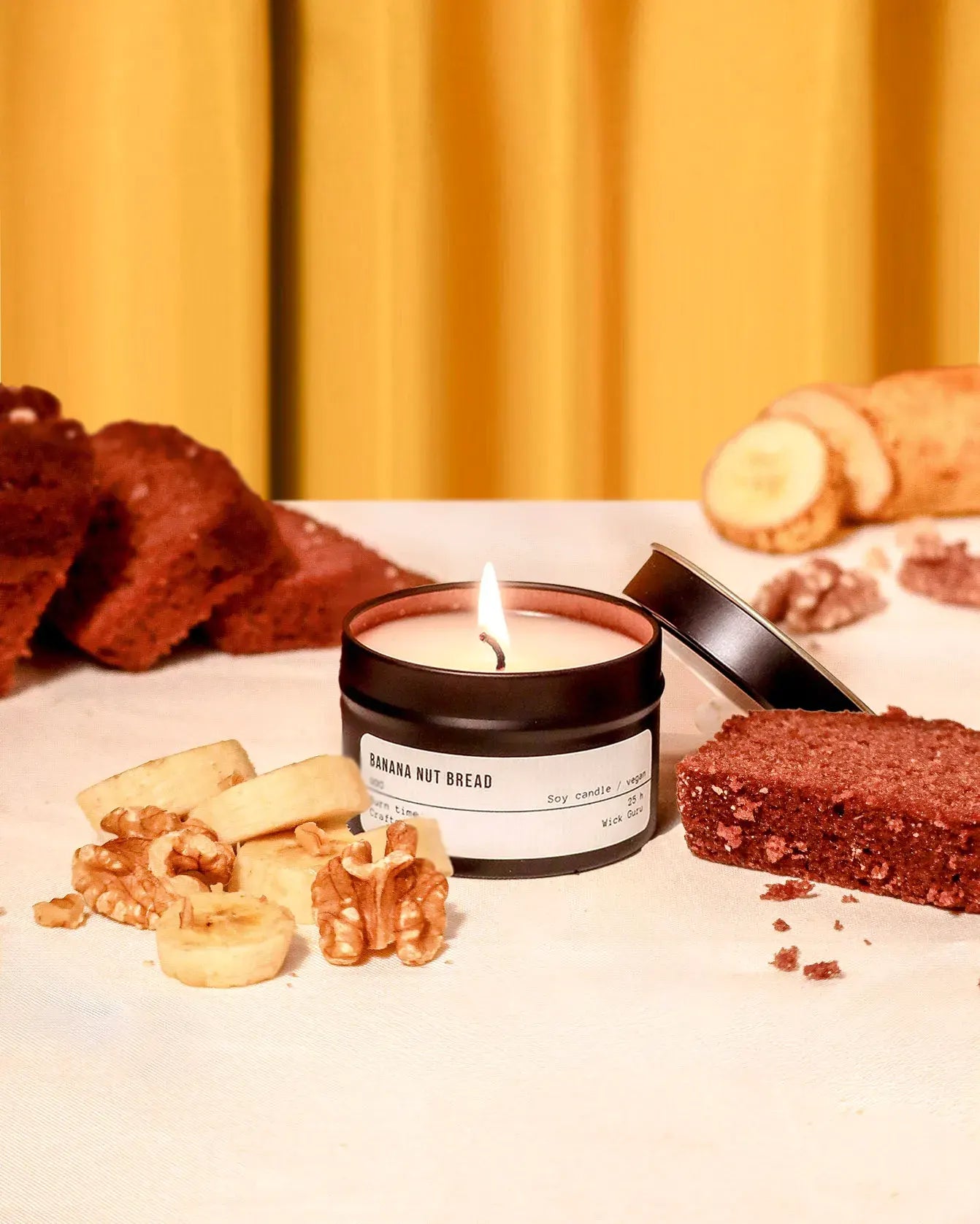 Candle labeled 'Banana Nut Bread' with banana, bread, and nuts on a table against a yellow curtain background.