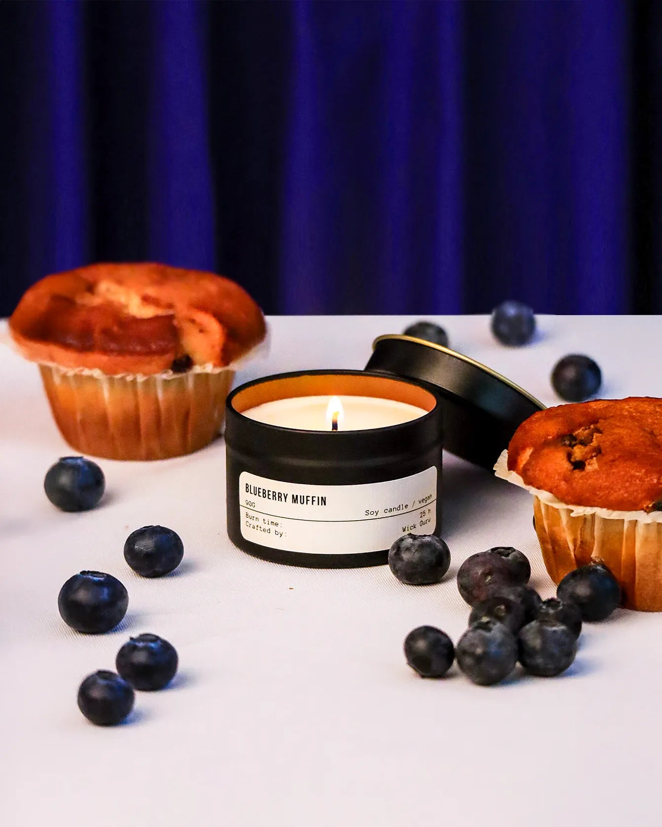 Candle labeled 'Blueberry Muffin' with blueberries and muffins on a white surface against a dark background