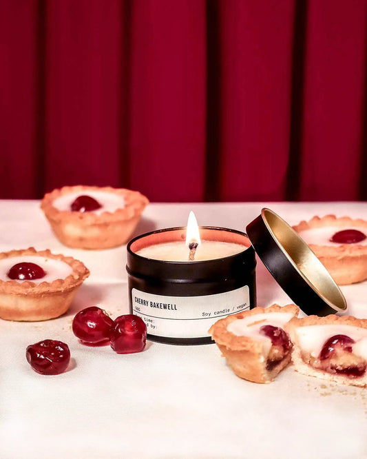 Candle labeled 'Cherry Bakewell' with cherry pies and cherries on a white surface against a red curtain background.