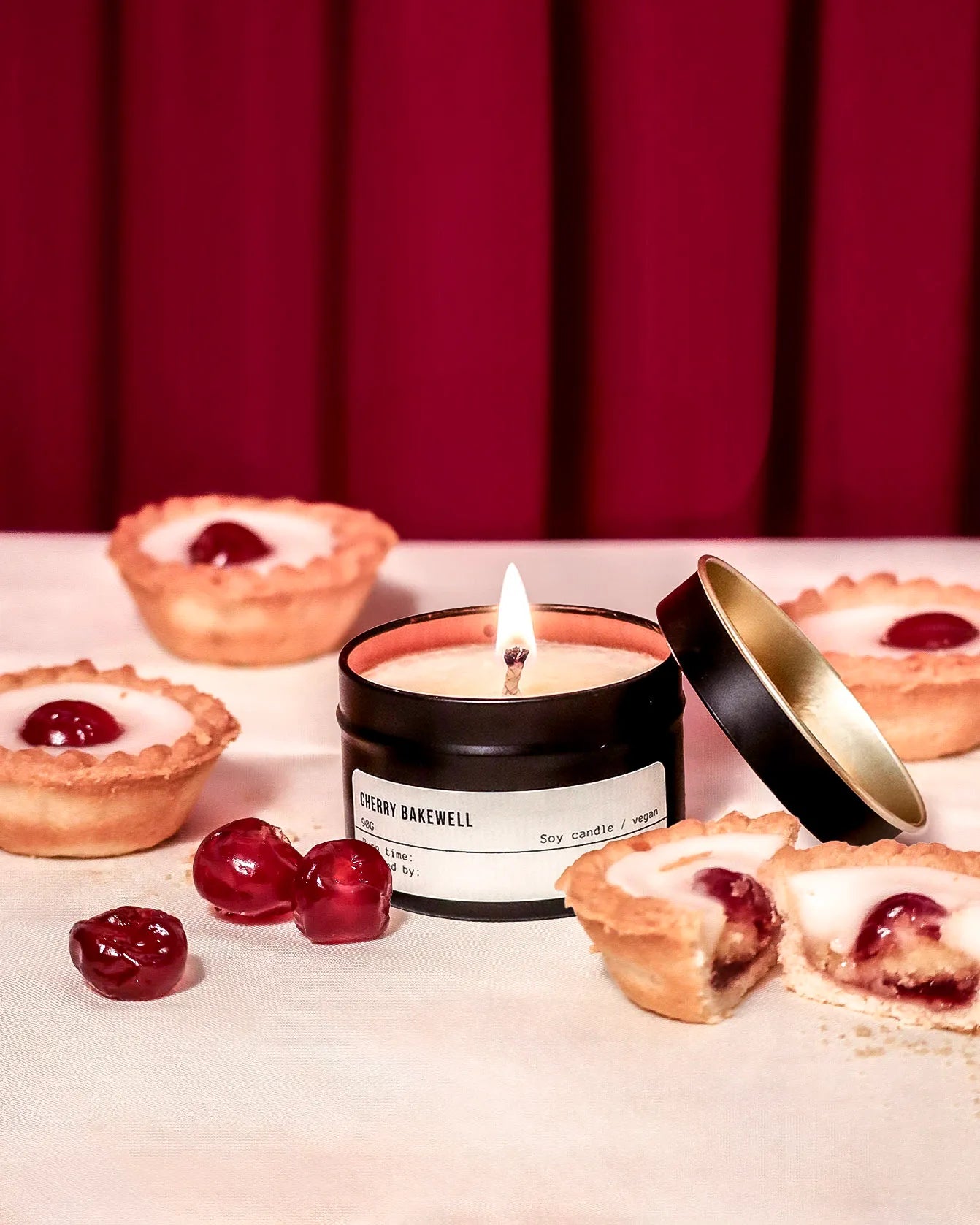Candle labeled 'Cherry Barewell' with cherry tartlets on a red curtain background