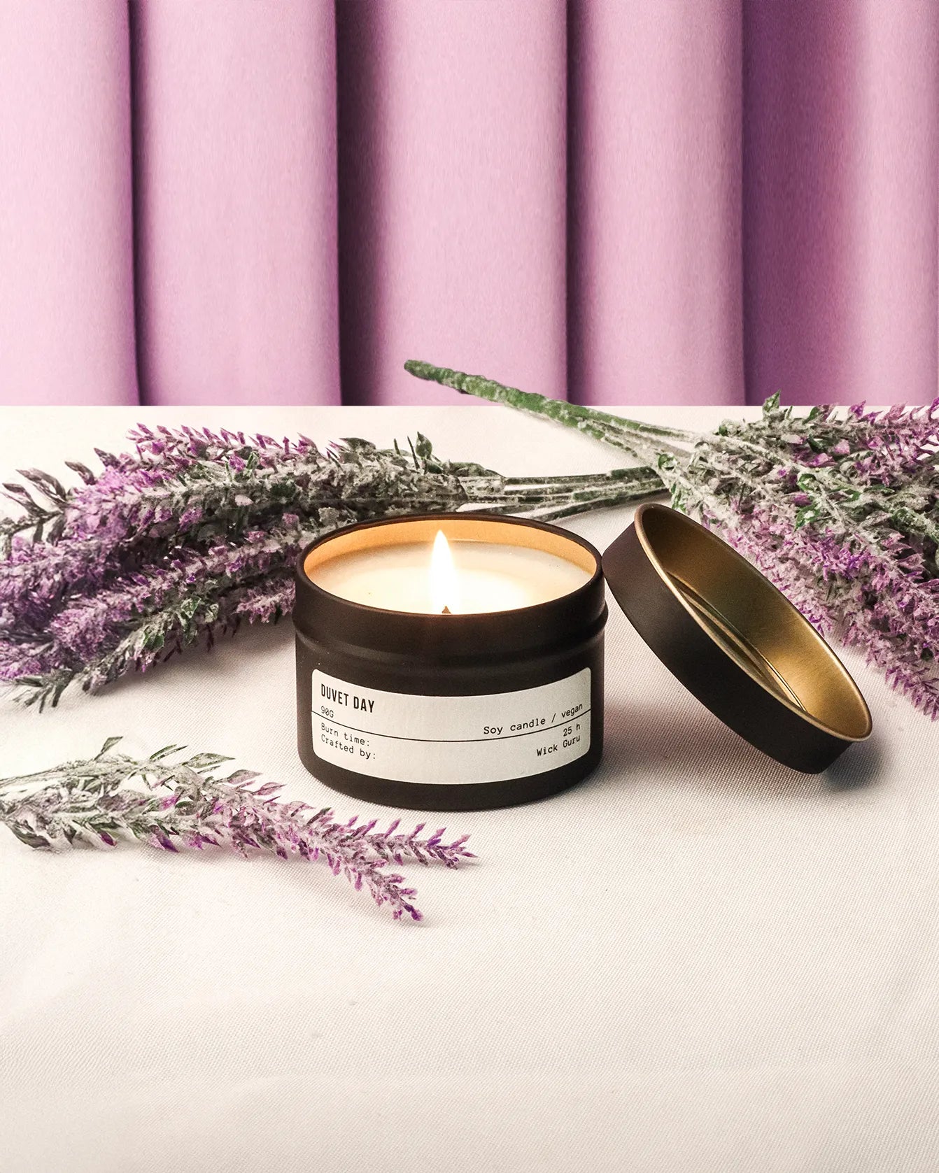 Candle in a black container with a gold lid, surrounded by lavender flowers against a purple curtain background.