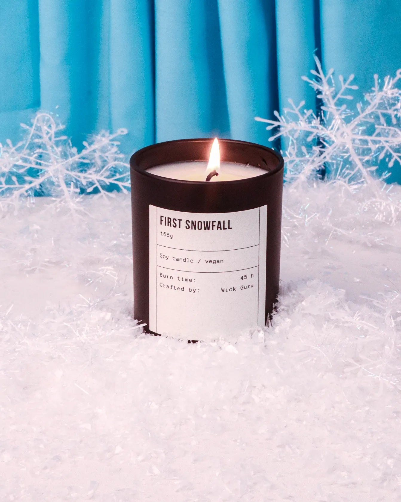 Candle labeled 'First Snowfall' in front of blue curtains with snowflake decorations