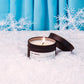 Candle labeled 'First Snowfall' in a snowy setting with blue curtains in the background