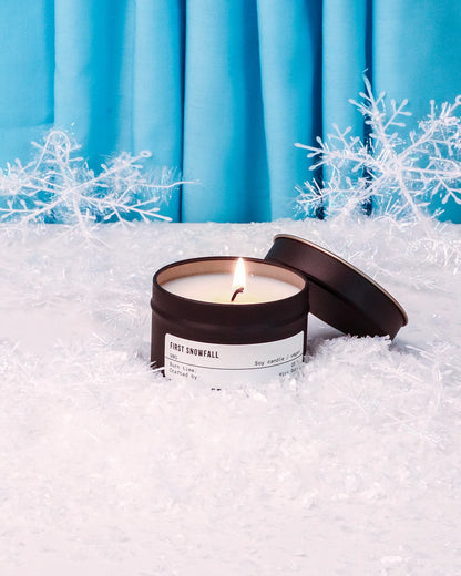 Candle labeled 'First Snowfall' in a snowy setting with blue curtains in the background