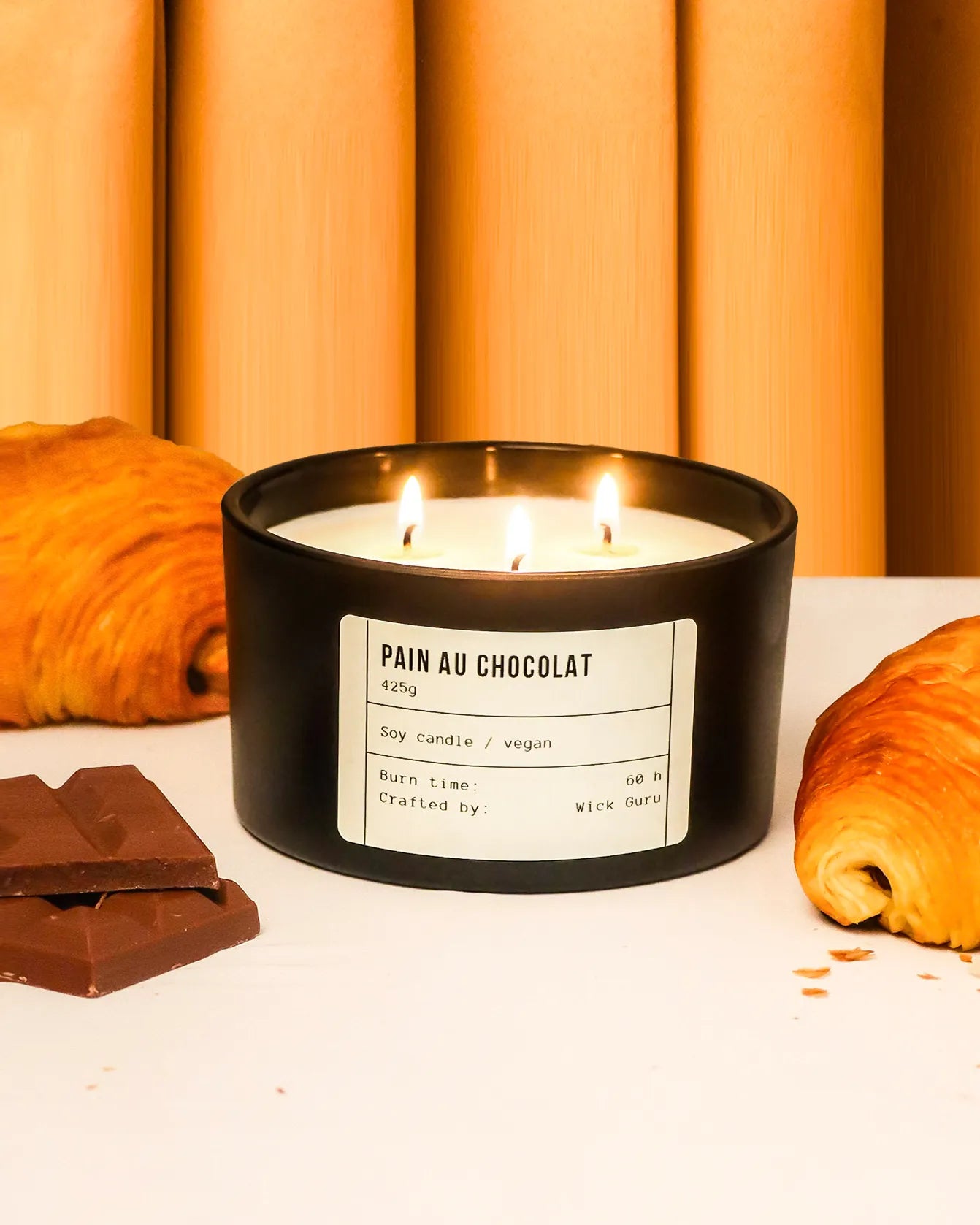 Candle labeled 'Pain au Chocolat' with croissants and chocolate bars on a white surface.