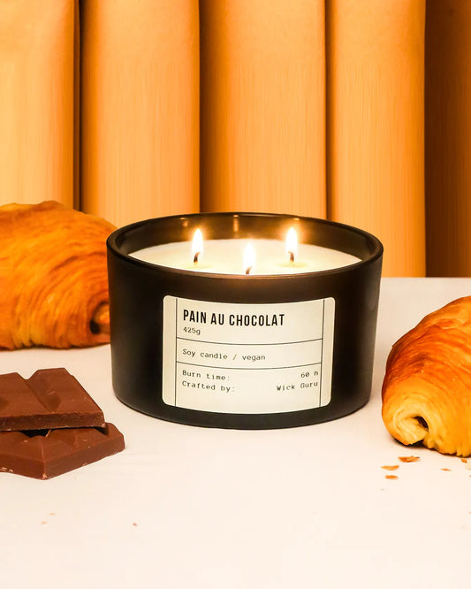 Candle labeled 'Pain au Chocolat' with croissants and chocolate bars on a white surface.