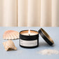 Candle in a black container with a white label, surrounded by shells on a light blue surface.