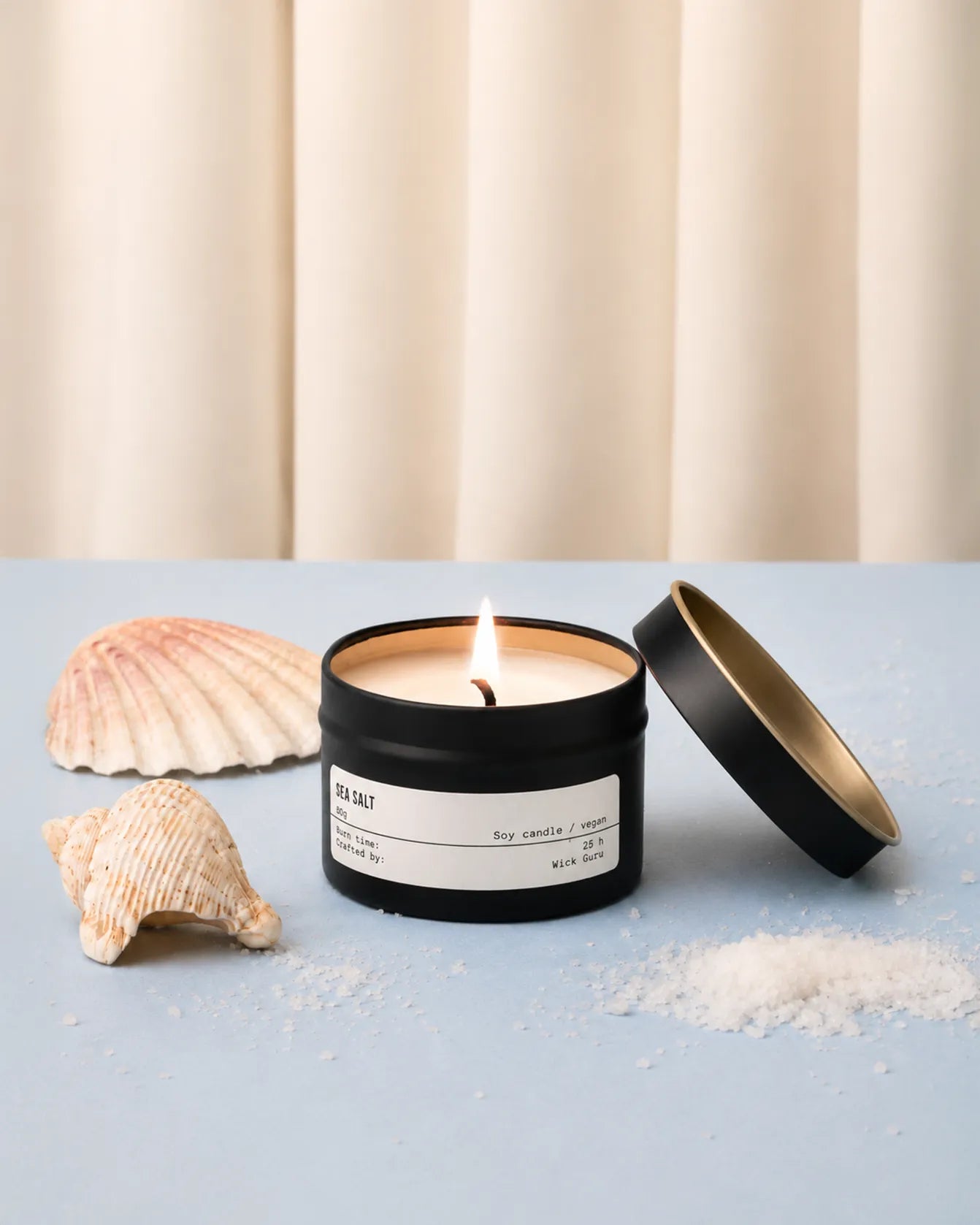Candle in a black container with a white label, surrounded by shells on a light blue surface.