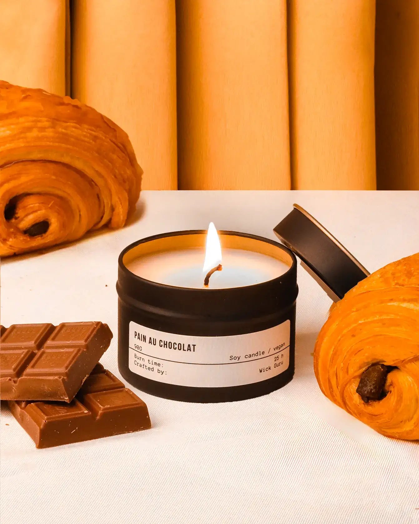 Candle labeled 'Pain au Chocolat' with chocolate bars and a croissant on a white surface.