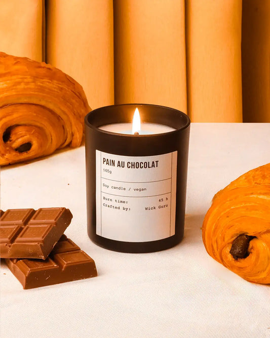 Candle labeled 'Pain au Chocolat' with chocolate bars and pastries on a white surface.