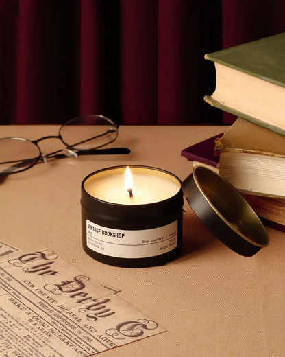 Candle in a black container labeled 'Vintage Bookshop' on a table with books and glasses.